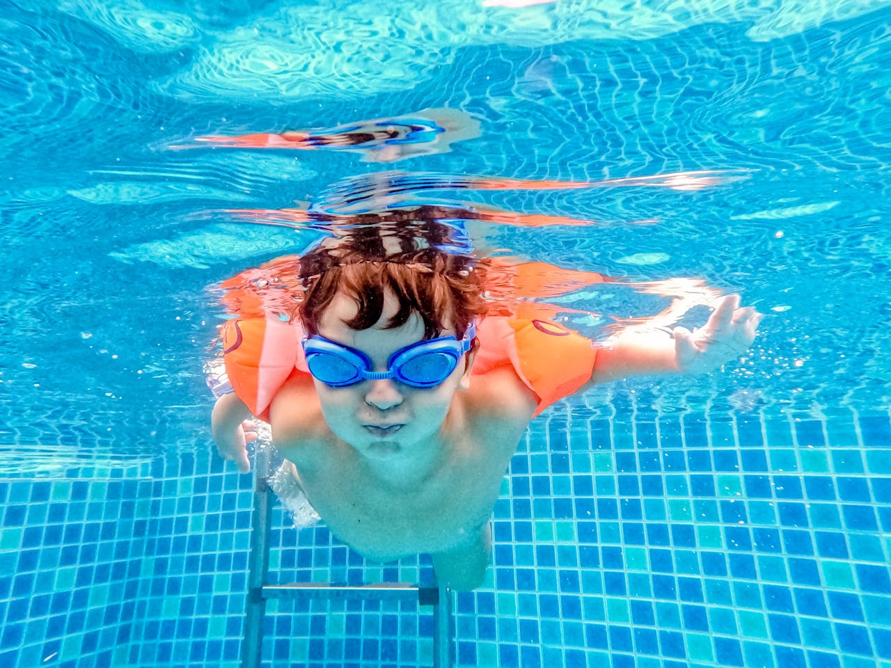 A child enjoying an underwater swim in a pool wearing goggles and arm floats.
